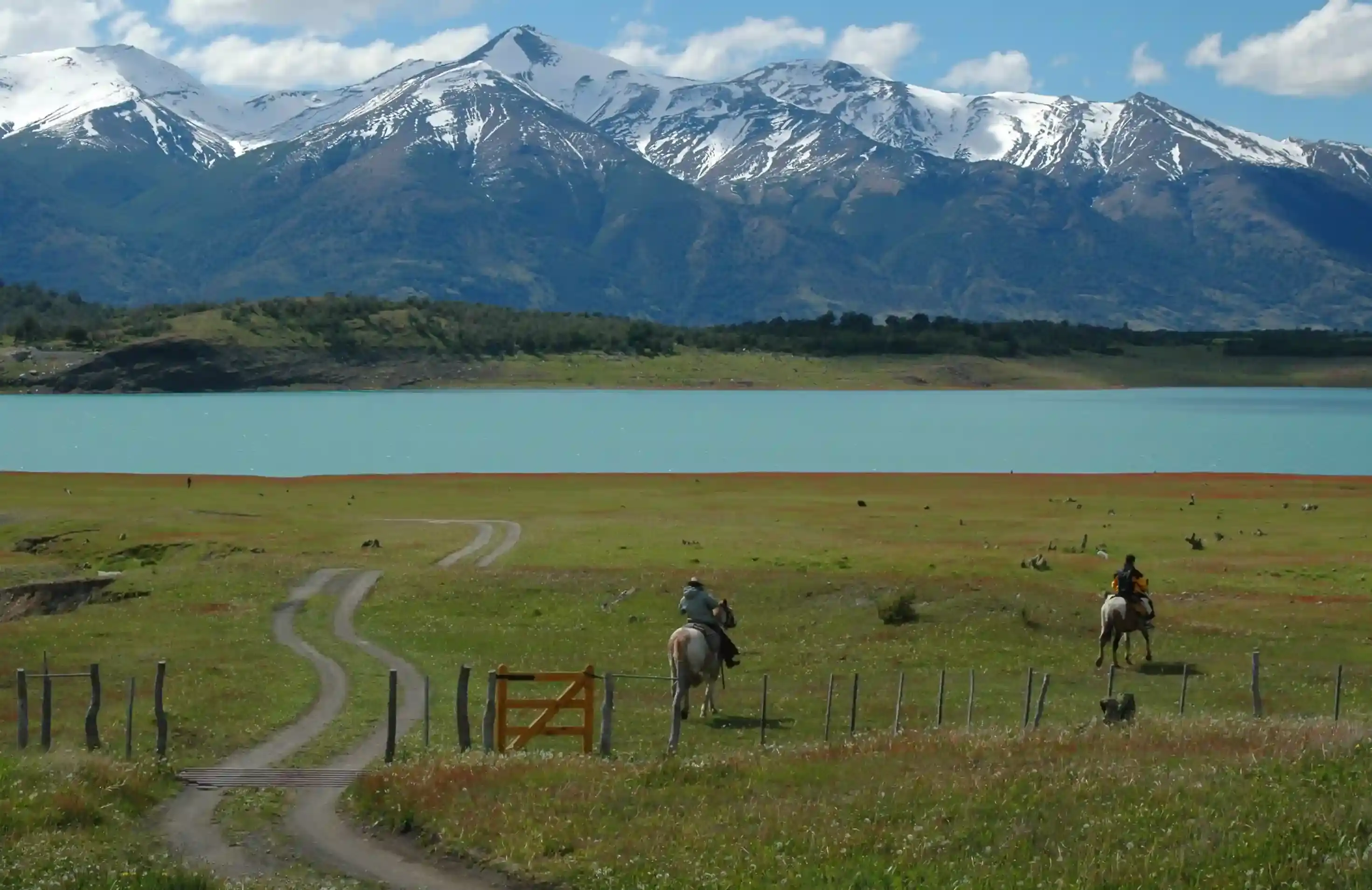 Gauchos on horseback by a turquoise lake under snowy Patagonian peaks